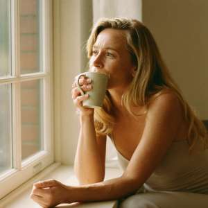A woman sitting by a sunlit window with a mug, looking calm and unhurried